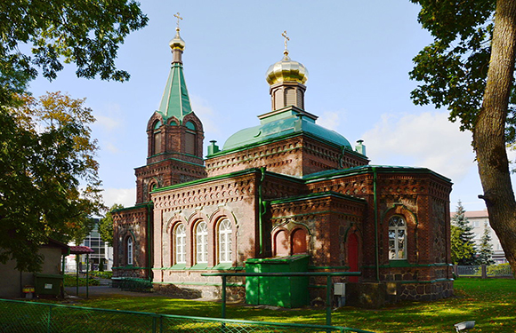 A small, ornate church with a green roof and a golden dome, surrounded by trees and a fence, under a clear sky.