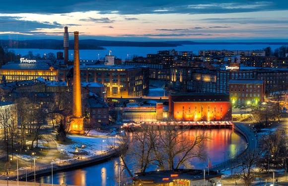 An image of a cityscape during twilight with illuminated buildings along a river, a prominent chimney structure, and a backdrop of a fading sky.