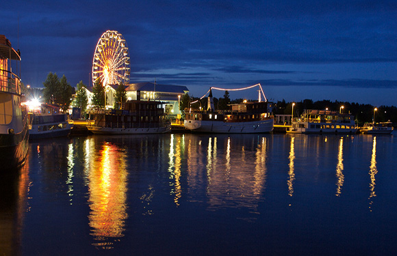 A nighttime waterfront scene with lit buildings, a Ferris wheel reflecting on water, and docked boats.