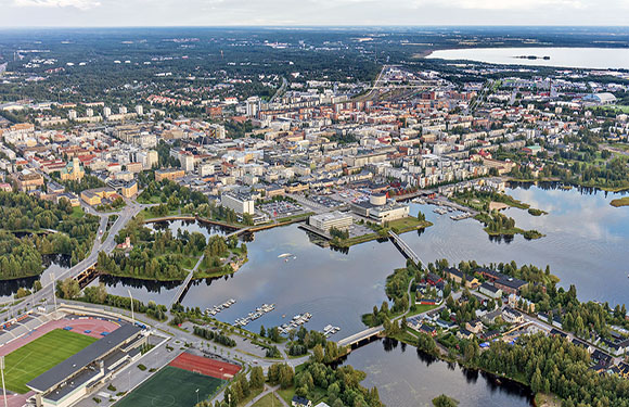 Aerial view of a town with buildings, a river with bridges, surrounded by greenery, water bodies, under a cloudy sky.