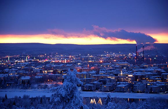 A twilight cityscape with snow, lit buildings, a smoking stack, under a purple-orange sky.