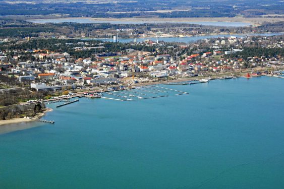 Aerial view of a coastal town with buildings near the shoreline, a pier extending into the blue water, and a backdrop of land and trees.