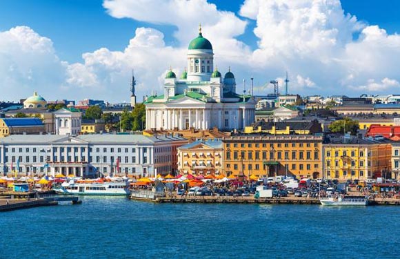 A coastal city with a white cathedral, twin bell towers, a bustling harbor with boats and tents, under a partly cloudy sky.