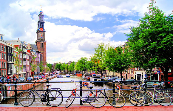 A lively city with a canal, boats, European buildings, parked bicycles, and a towering structure.