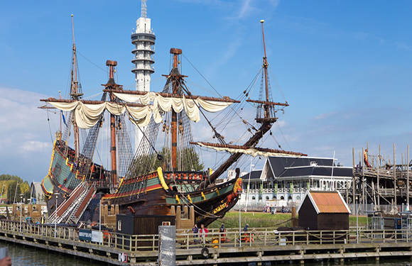 A historical tall ship with multiple masts and sails docked at a pier, with a clear sky above and modern buildings in the background.