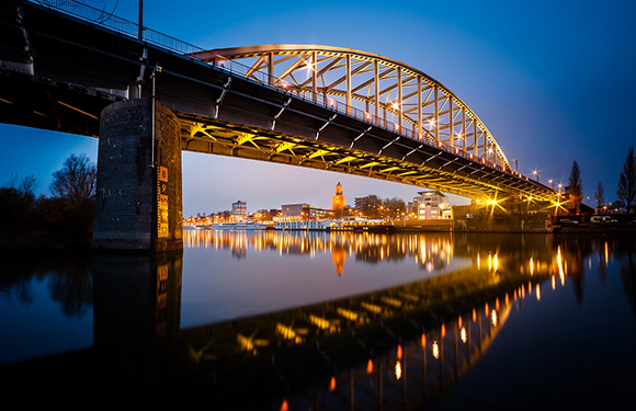 An image of a lit bridge over a calm river during twilight, with lights reflecting on the water and a town visible in the background.