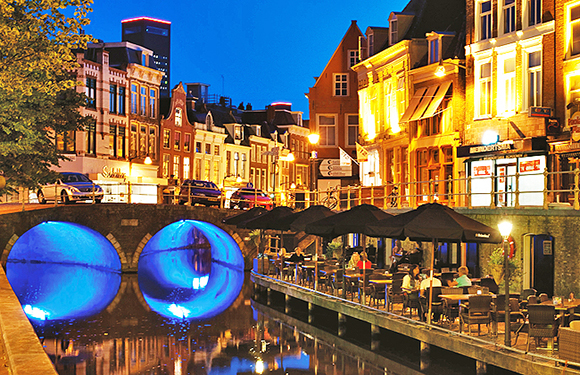 A vibrant evening scene of a city canal with illuminated buildings and bridges, outdoor seating at restaurants, and reflections on the water.