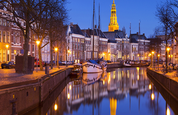 A nighttime cityscape with illuminated buildings and a church spire reflected in a calm canal, flanked by moored boats and parked cars.