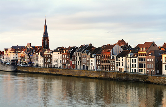 A row of traditional European buildings along a riverbank with a clear sky above and a church spire in the distance.