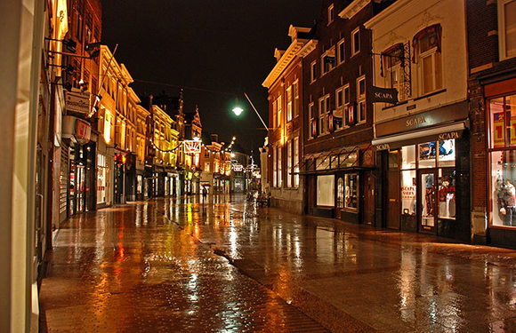 A wet street at night reflecting city lights, with buildings on either side and no visible people.