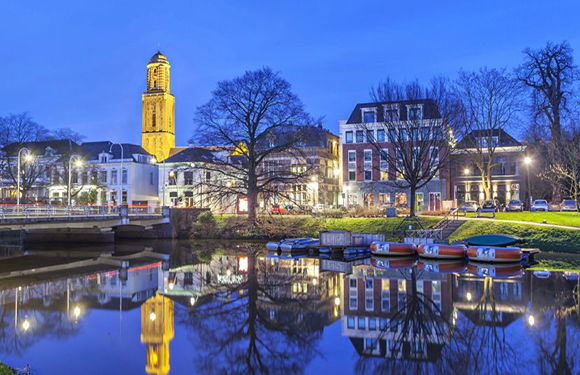 A scenic evening view of a calm river with reflections of surrounding buildings and boats, and a prominent tower in the background.