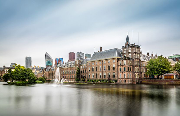 A historic building with towers and spires by a fountain and water, with modern skyscrapers under a cloudy sky in the background.