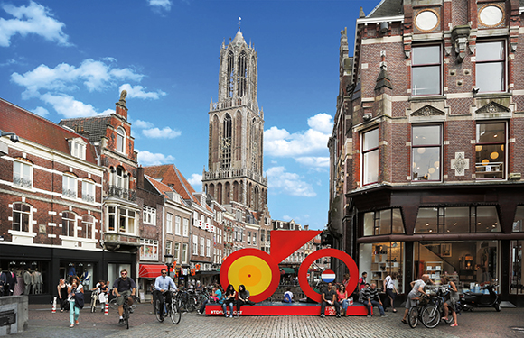 A European street scene with people, traditional buildings, a large abstract sculpture, and a Gothic church tower.