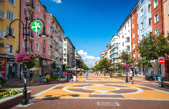 A lively street with colorful buildings, people, decorative lamps, flowers, a circular pavement pattern, and a no entry sign.