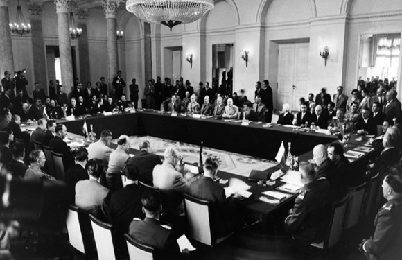 Black and white image of a formal meeting with many participants, seated and standing, in a chandelier-lit room.