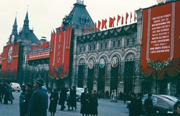 A historical color photo showing a street scene with people walking by a large building adorned with red banners and flags.