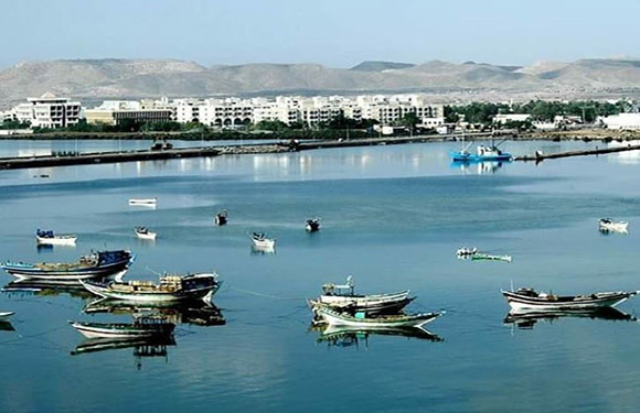 A coastal scene with multiple boats floating on calm water, with buildings in the background against a backdrop of hills or mountains.