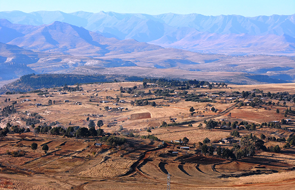 A landscape view of a rural area with scattered buildings, fields, and dirt roads, with mountains in the background under a clear sky.