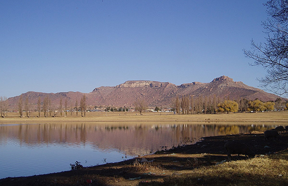 A tranquil landscape featuring a calm lake with reflections of a mountain range and trees in the background under a clear blue sky.