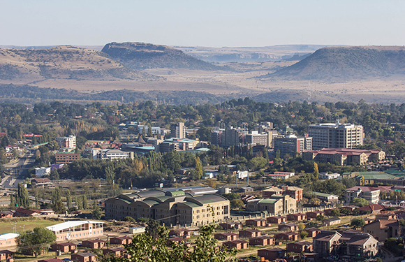 A panoramic view of a city with buildings of various sizes, surrounded by a hilly landscape in the background.