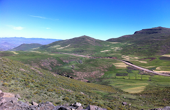 A scenic landscape featuring rolling hills with patches of green vegetation and cultivated fields under a clear blue sky.