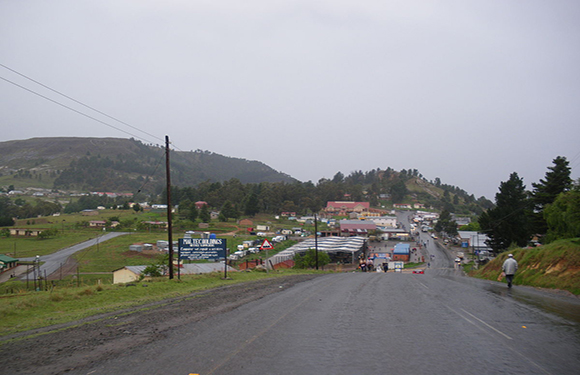 A person walks away on a road into a small town under a cloudy sky, flanked by buildings and trees.