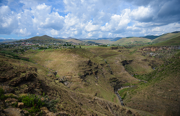 A picturesque scene of rolling hills, sparse vegetation, a stream, and a distant settlement under a partly cloudy sky.