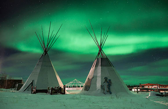 A photo shows two teepees under the Northern Lights with a person's silhouette, distant lights and snowy ground.