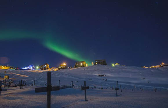 A night-time scene with the Northern Lights (aurora borealis) visible in the sky, over a snowy landscape with illuminated buildings.