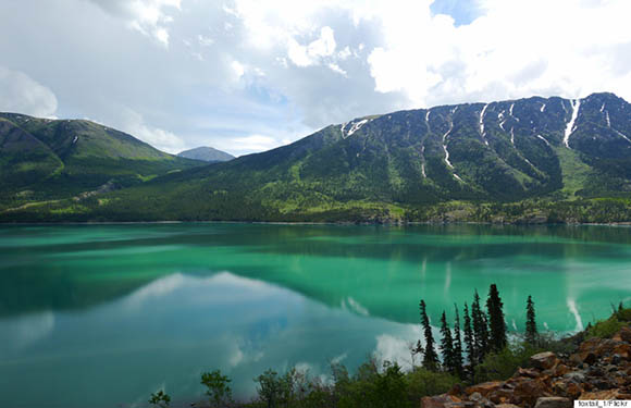 A scenic view of a tranquil turquoise lake with reflections of surrounding mountains and clouds in the water, under a partly cloudy sky.