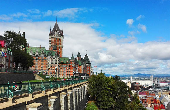 A historic castle-like building with a tall tower overlooks a cityscape and river under a partly cloudy sky.