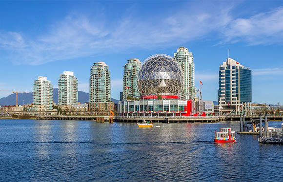 A city waterfront features modern skyscrapers, a unique geodesic dome, and a small red and white boat under a clear blue sky.