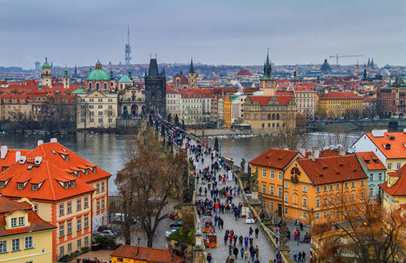 Aerial view of a cityscape with historical buildings, a bridge crowded with people, and a river running through the center, under an overcast sky.