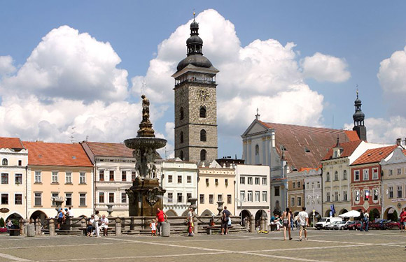 A town square features a fountain, tower, historic buildings, and people under a partly cloudy sky.