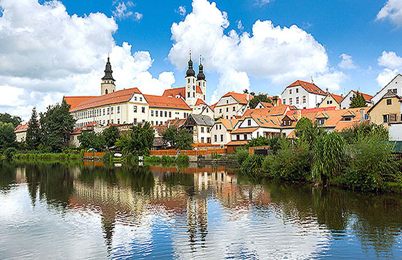 A picturesque view of a European town with historic buildings and spires reflected in a calm river under a partly cloudy sky.