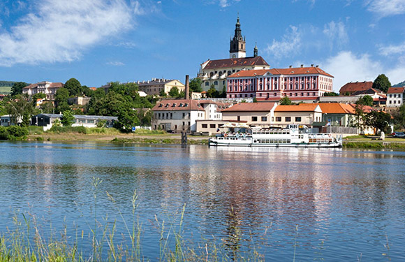 A picturesque river scene with a docked boat, historic town, prominent church spire, and a blue, partly cloudy sky.