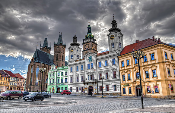 A scenic European town square with baroque and renaissance buildings, parked cars, and no people under a cloudy sky.