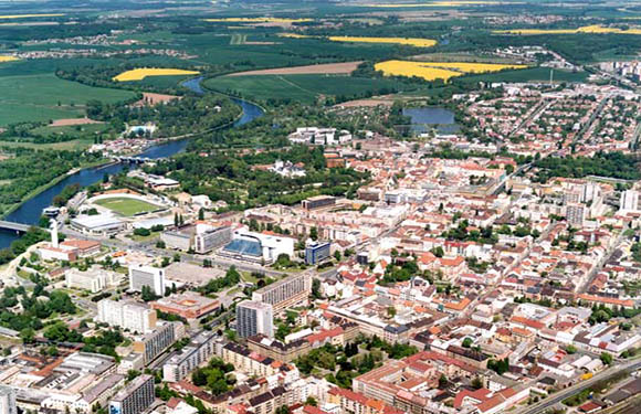 Aerial view of a town with buildings, streets, and greenery, intersected by a river.