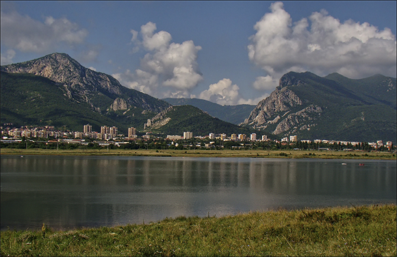 A scenic view of a calm lake with a cityscape in the background, nestled at the foot of a mountain range under a blue sky with scattered clouds.