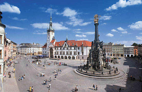 A lively town square with a central statue, surrounded by classic European architecture under a partly cloudy sky.