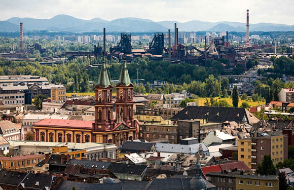 Aerial cityscape view featuring a church with green spires, industrial buildings, mountains, and a cloudy sky.