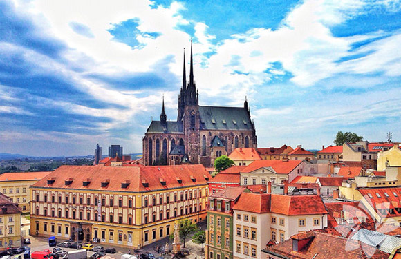 Aerial view of a European cityscape with Gothic cathedral spires rising above orange-roofed buildings under a partly cloudy sky.