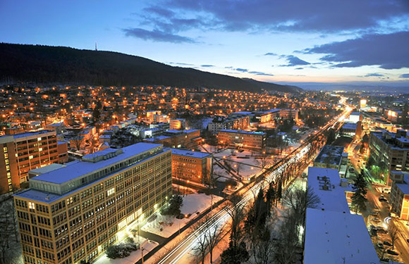 A cityscape during twilight with illuminated streets and buildings, snow-covered areas, and a mountain in the background.