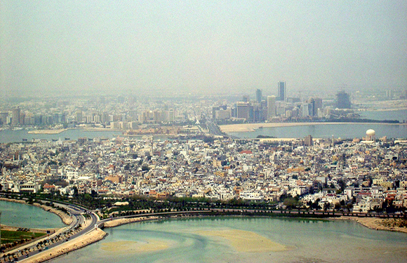 Aerial view of a crowded city with nearby river and distant coastline, under a hazy sky with earthy colors.