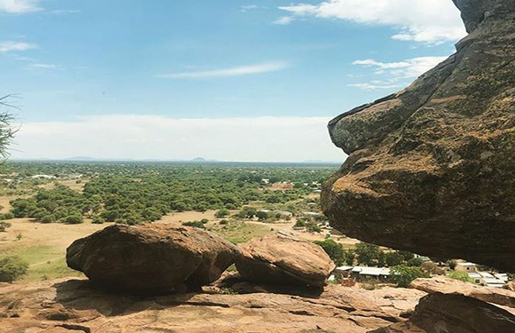 A high vantage point offers a scenic view of a vast landscape with scattered trees, large rocks, and a partly cloudy sky.