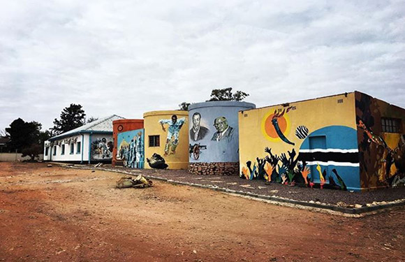 A row of colorful buildings with vibrant murals painted on their exterior walls, depicting various figures and patterns, under a cloudy sky.