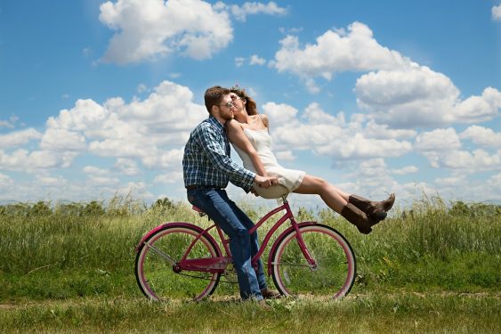 A man and a woman embracing on a pink bicycle in a grassy field with a blue sky and clouds in the background.