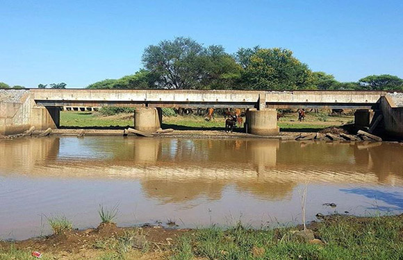 A concrete bridge over a body of water with reflections visible on the surface, surrounded by greenery and clear skies.