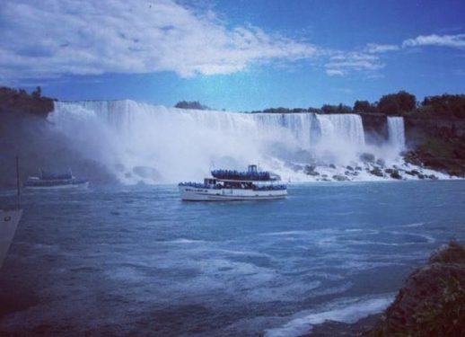 A boat with tourists approaches a large waterfall under a partly cloudy sky.