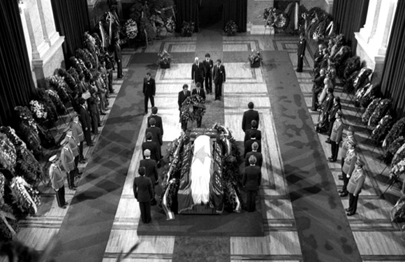 A monochrome photo shows a solemn procession in a hall, with a coffin carried by people, surrounded by attentive military personnel.